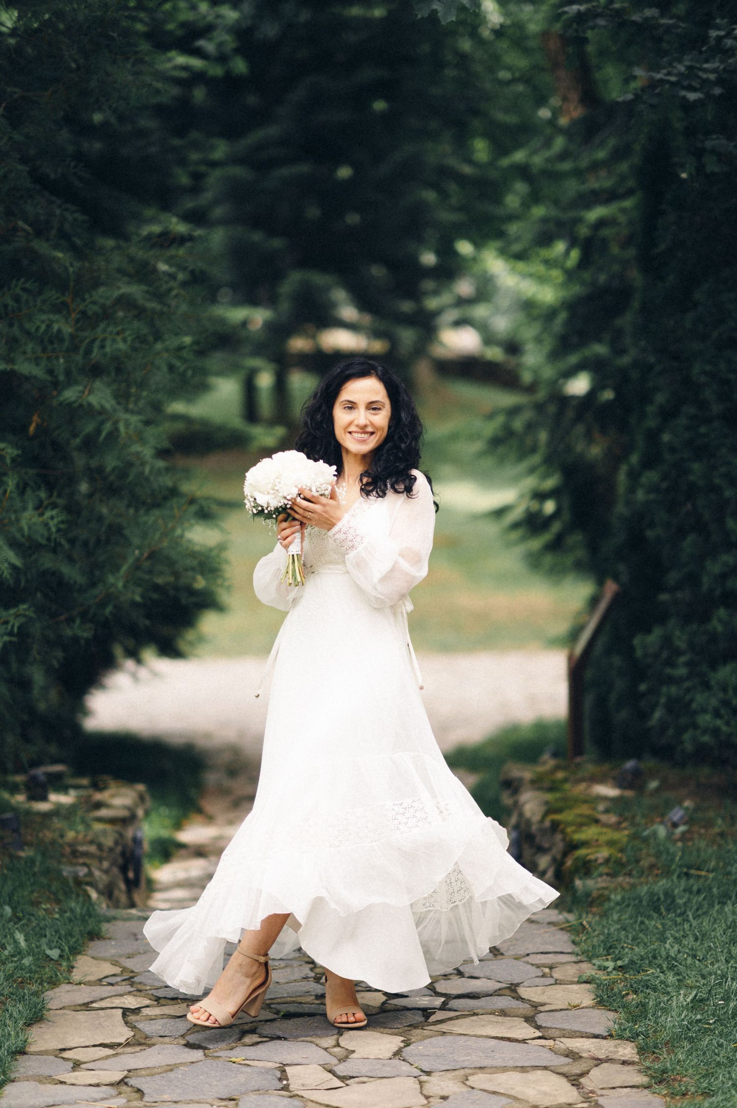 Bride in flowing white dress twirls on a stone pathway surrounded by greenery.