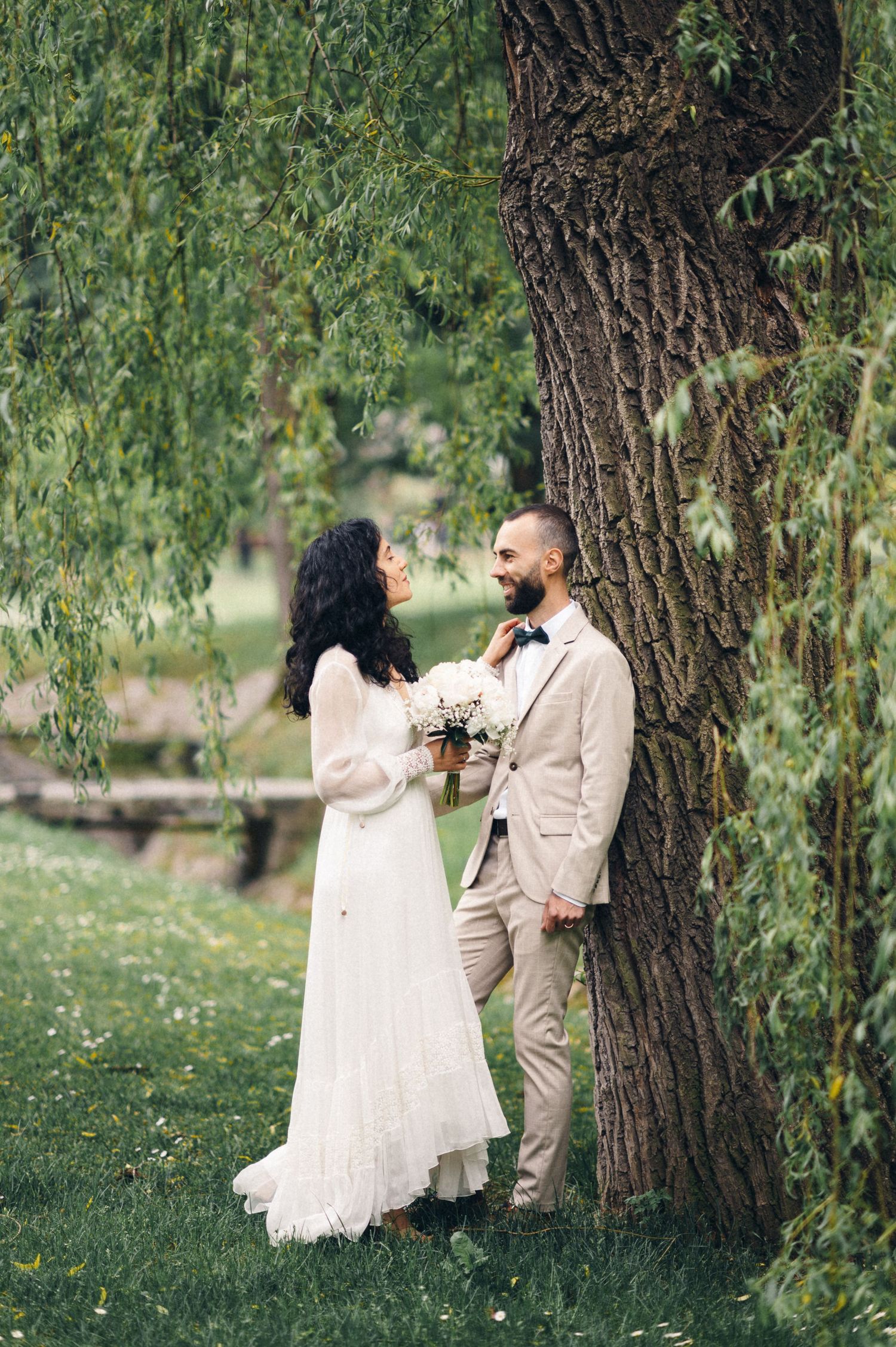 Newlyweds share a romantic moment under a willow tree in a garden setting.