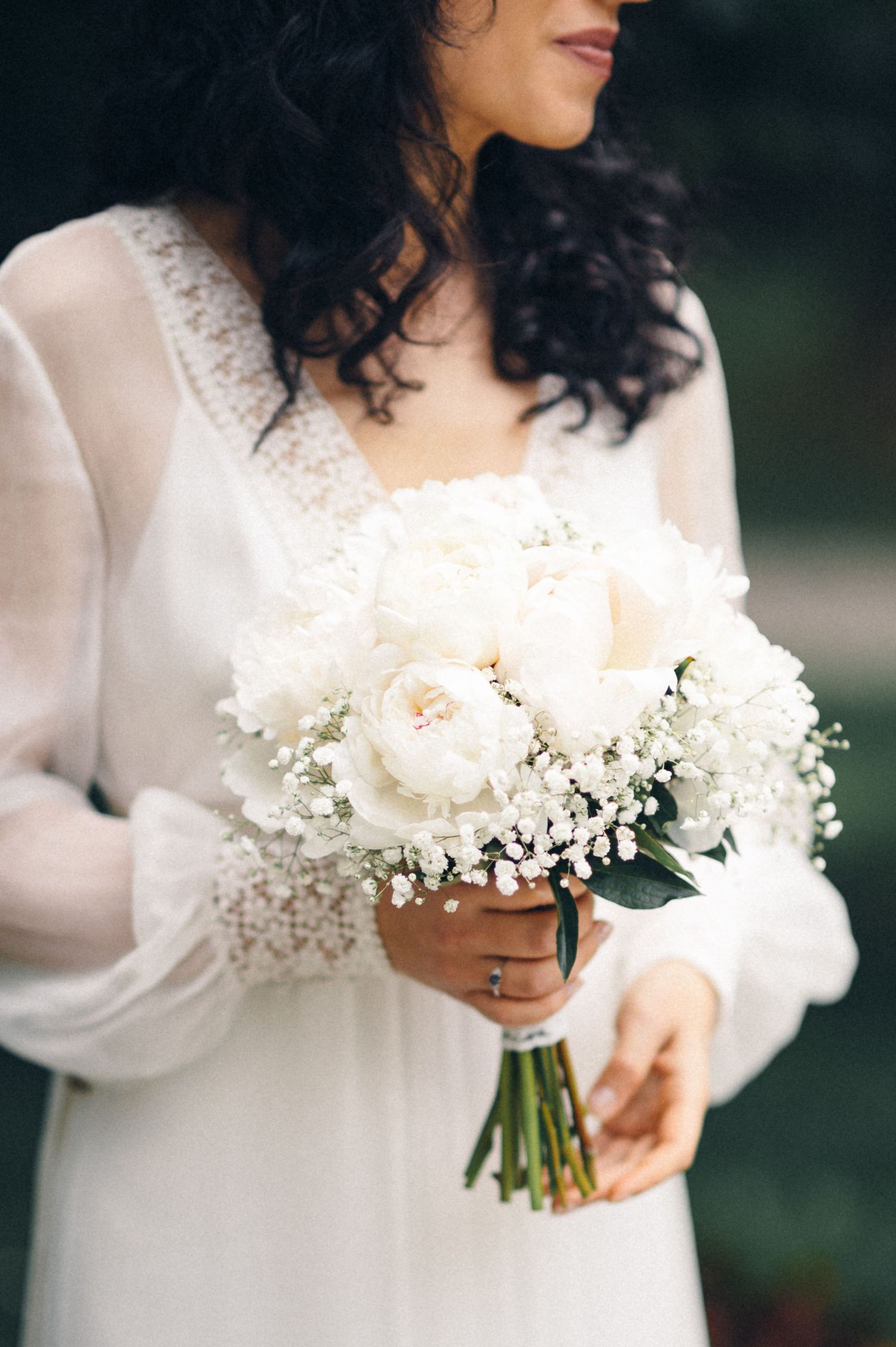 A delicate white bridal bouquet with roses and baby's breath held against a long-sleeved lace wedding dress.