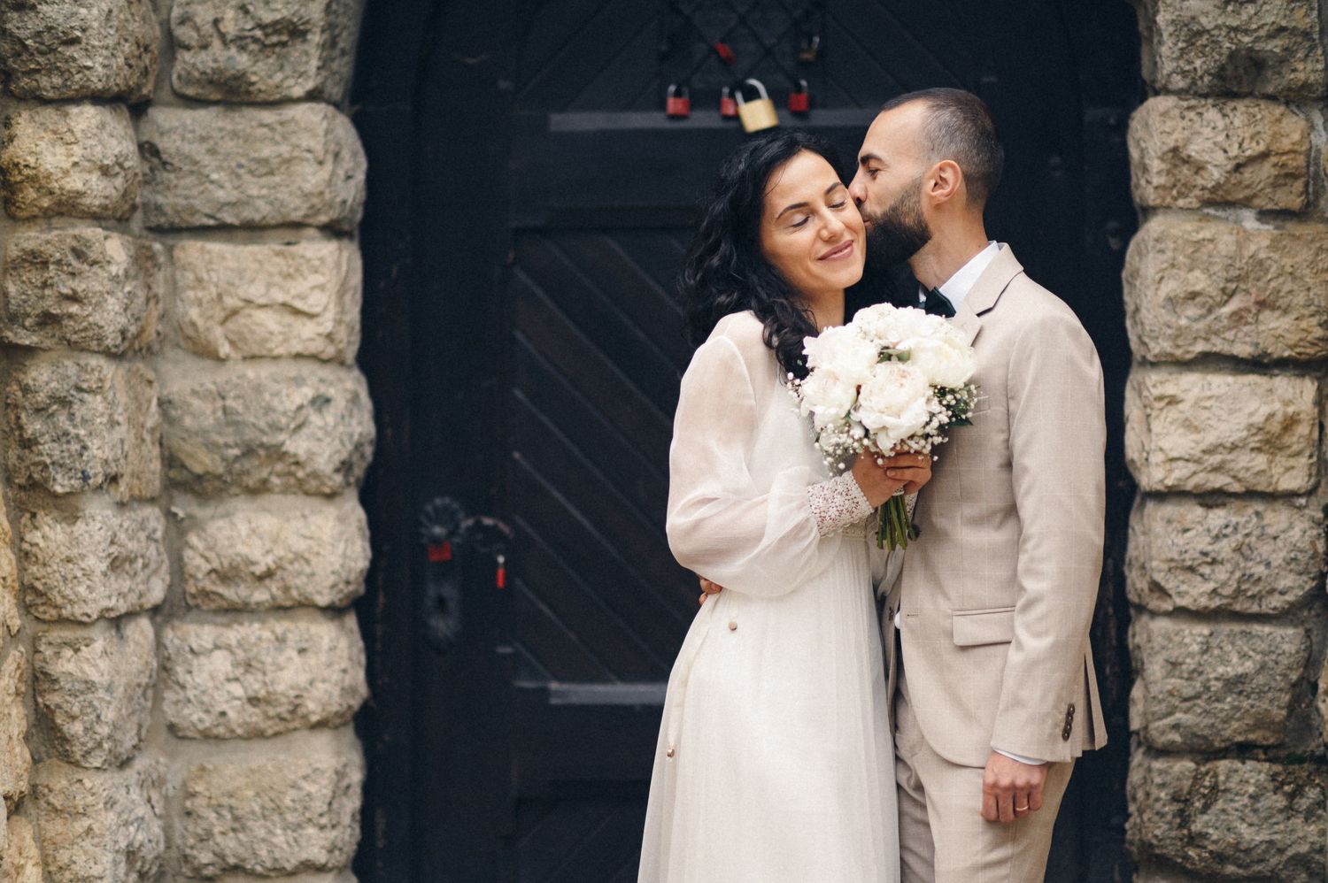 Couple in wedding attire embracing in front of a stone archway with vintage wooden door.