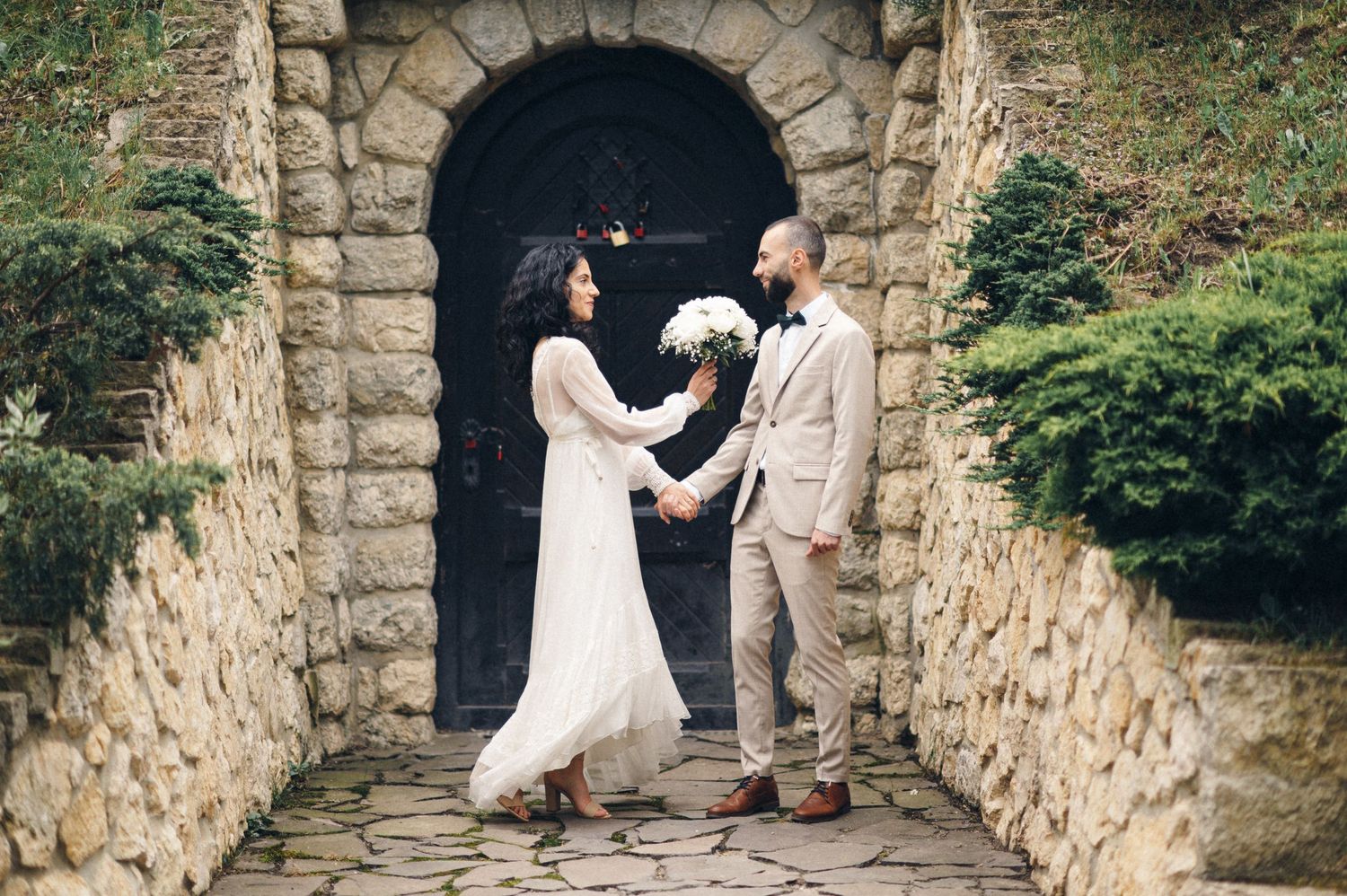 Couple in wedding attire holds hands beneath a rustic stone archway in a garden setting.