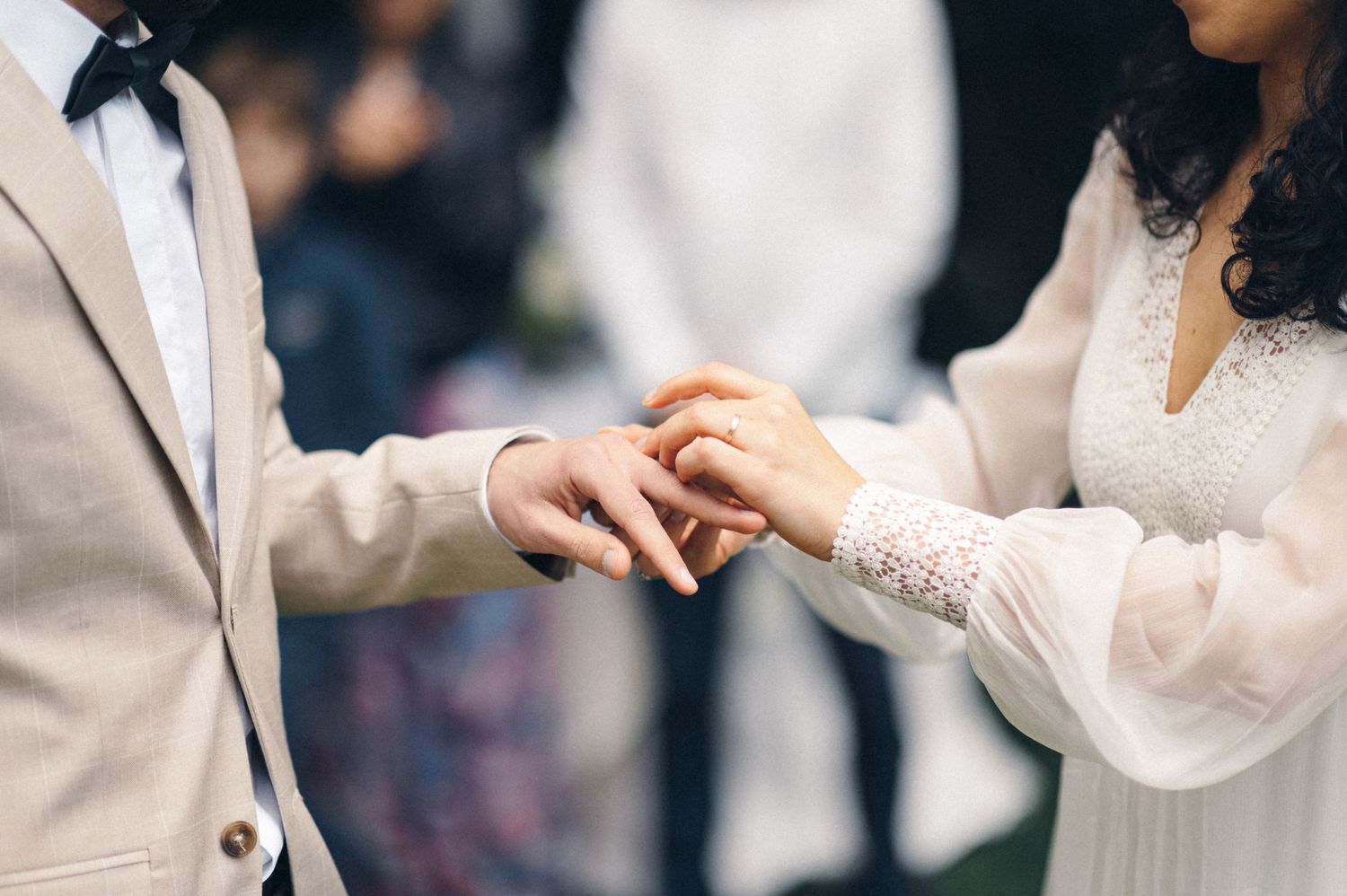 Close-up of hands exchanging wedding rings against white lace and beige suit sleeves.
