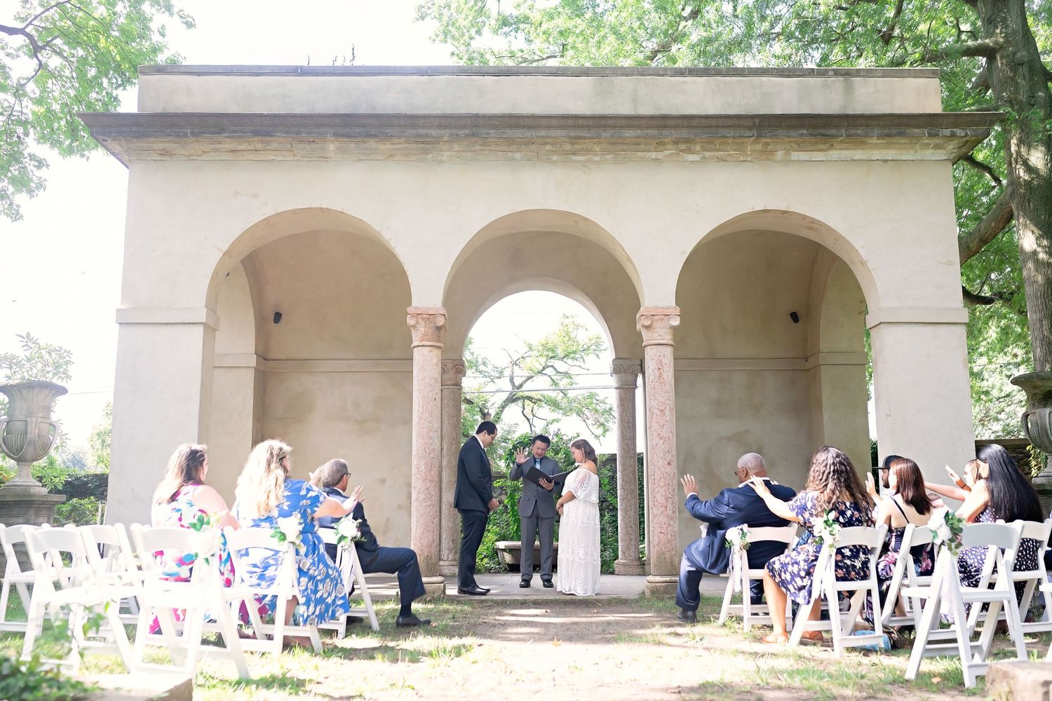 Small intimate outdoor wedding ceremony under stone archway with guests seated in white chairs.