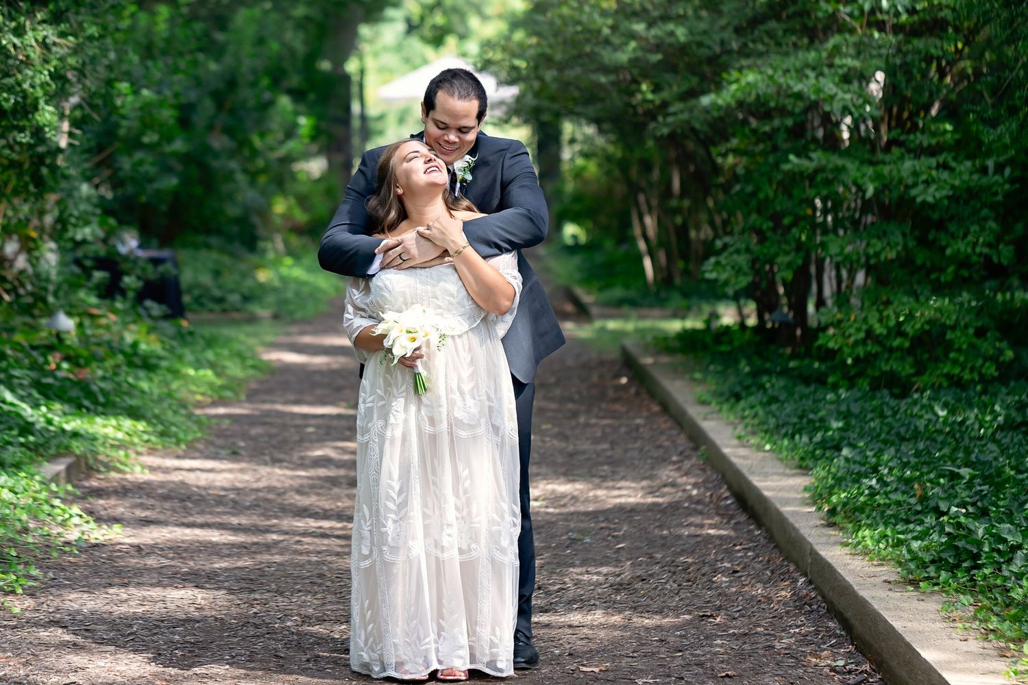 Groom embracing bride from behind on a tree-lined garden path as she holds white bouquet.
