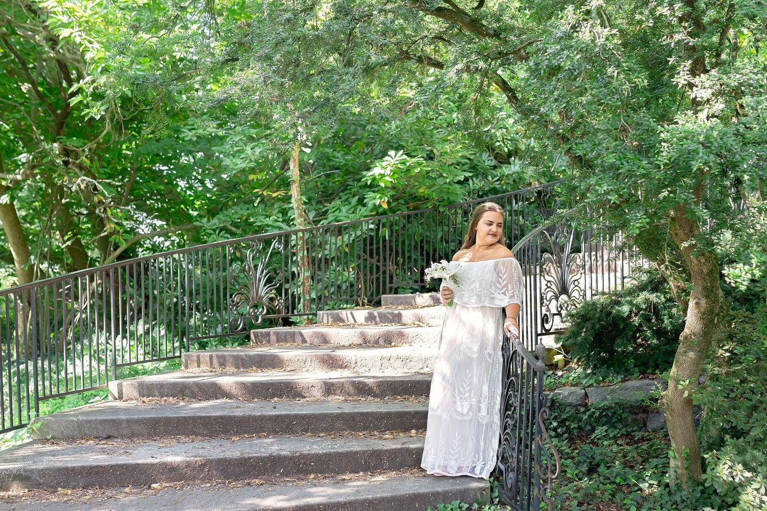 Bride in white lace off-shoulder gown holding bouquet on stone steps surrounded by lush greenery.