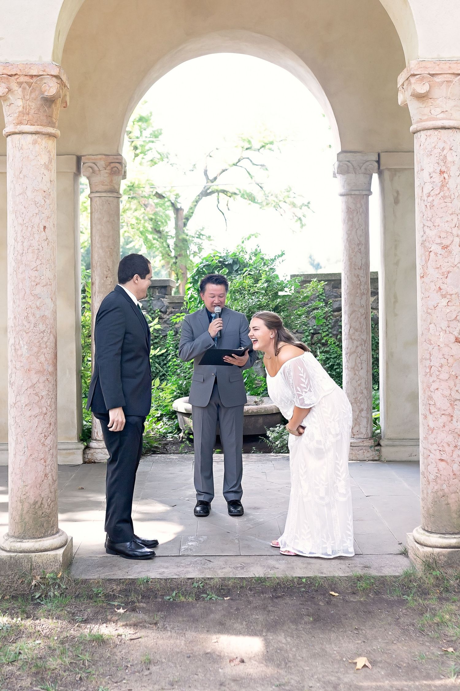 Bride and groom laughing with officiant during outdoor wedding ceremony under elegant stone arches.
