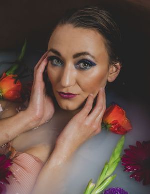 Woman with blue eye makeup and pink lips poses in a floral milk bath with orange roses and purple flowers.