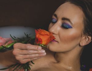 Woman with colorful eye makeup smells an orange rose in a floral bath setting.