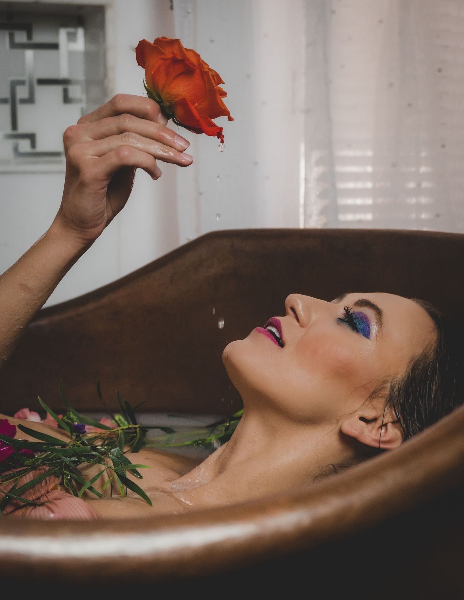 Woman in copper bathtub holding an orange rose with dramatic blue eye makeup and wet hair.