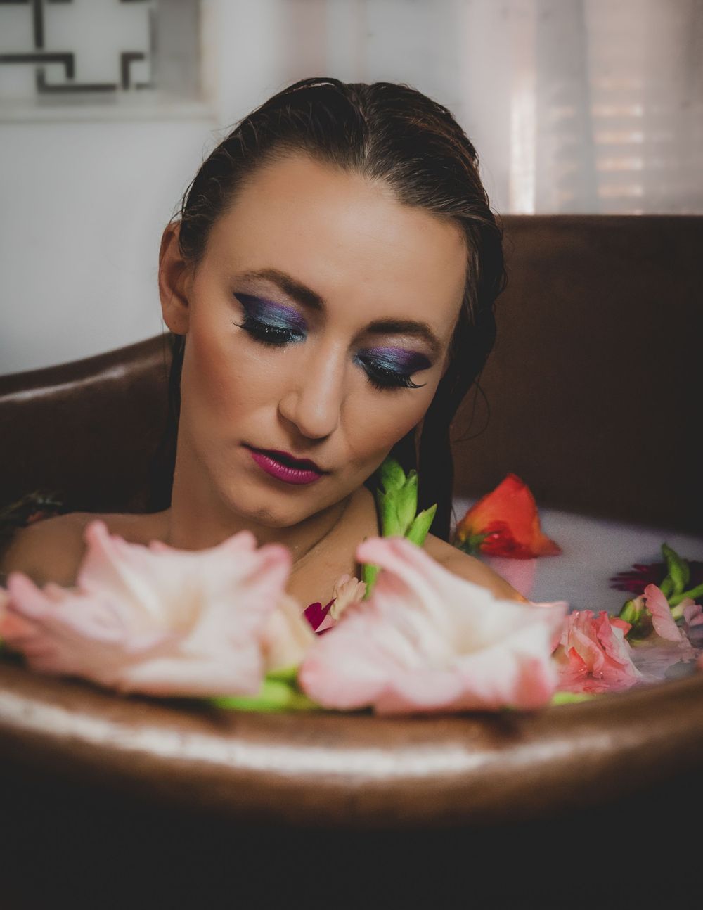 Woman with bold purple eye makeup gazing down at pink flowers in a copper milk bath.