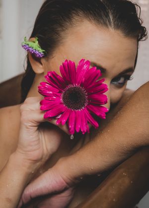 Woman holds a vibrant pink gerbera daisy over her face with a small purple flower in her hair.