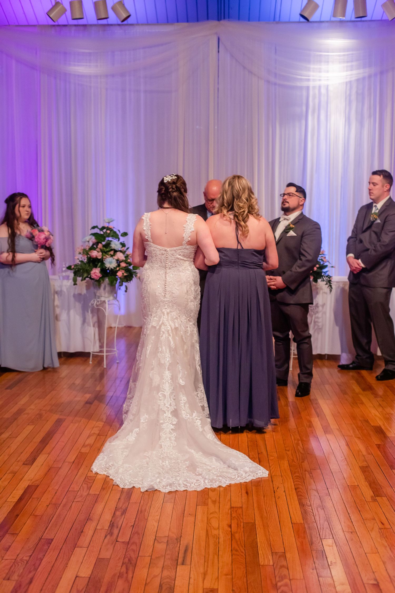 Wedding ceremony in a purple-lit venue with elegant white drapes and hardwood floors.