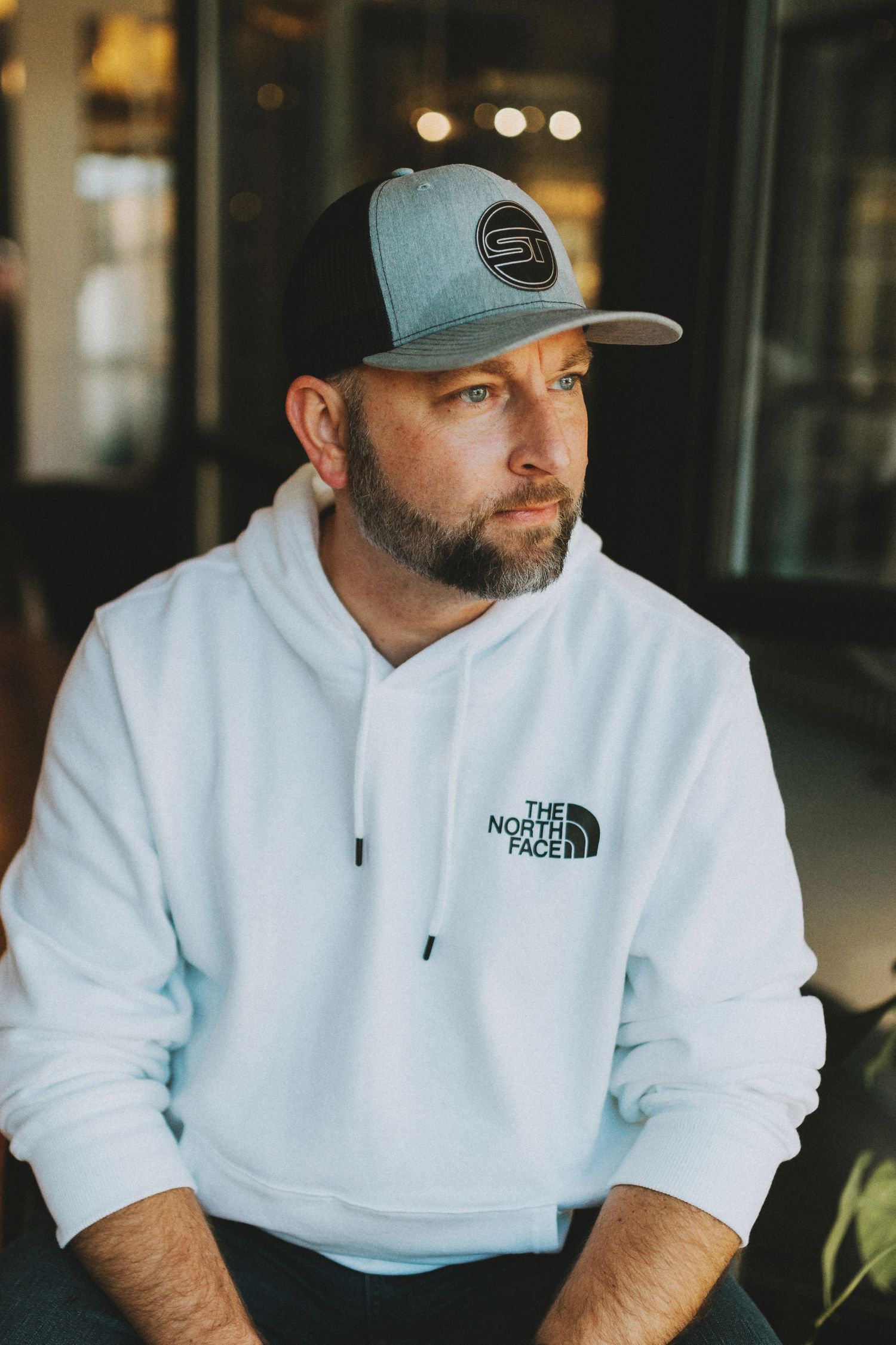 Casual portrait in white North Face hoodie and teal cap with soft indoor lighting.