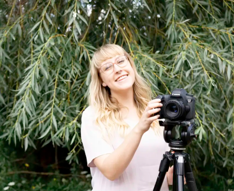 Photographer smiling while using camera on tripod among willow trees.