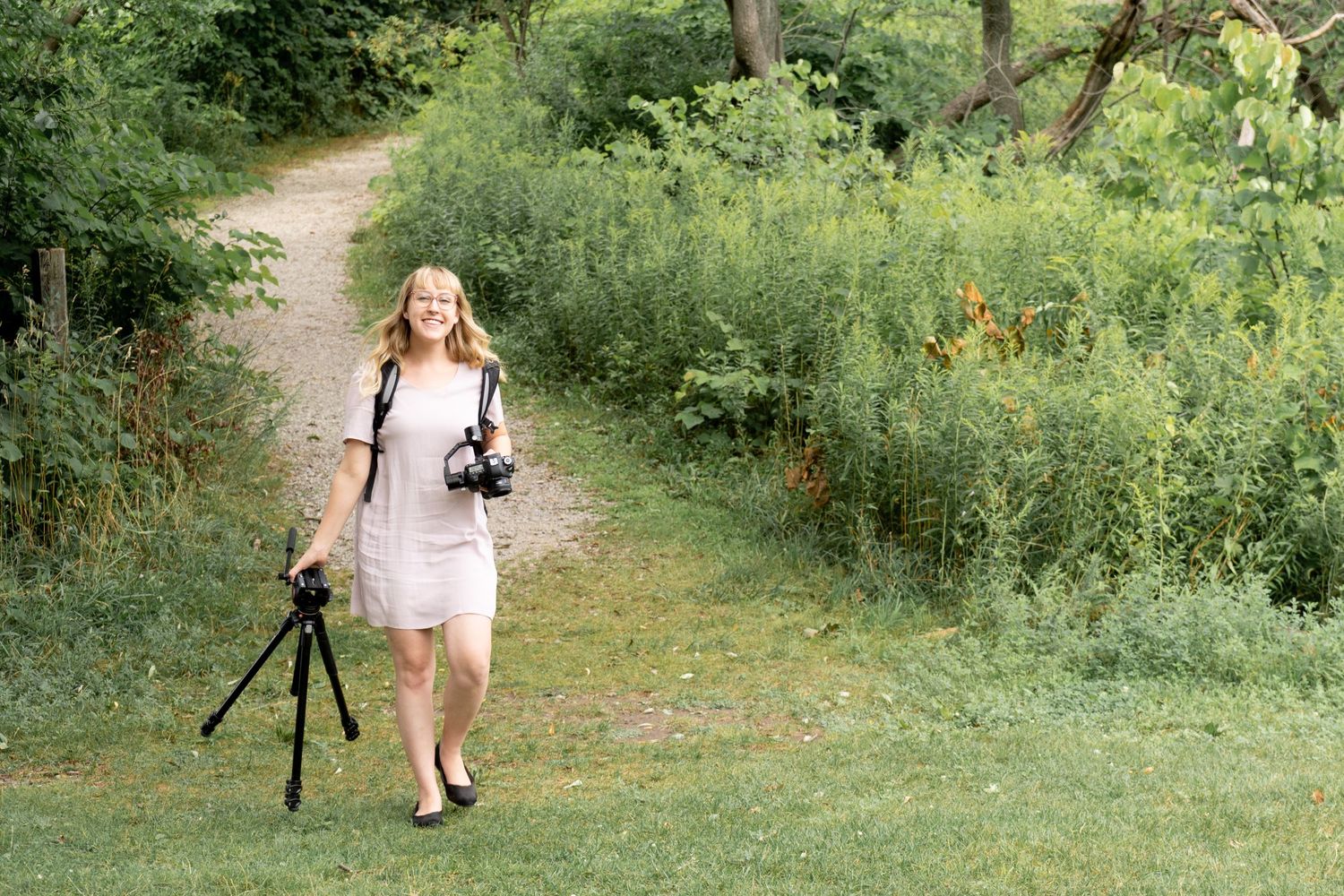 Photographer walking on nature trail carrying camera and tripod in summer dress.