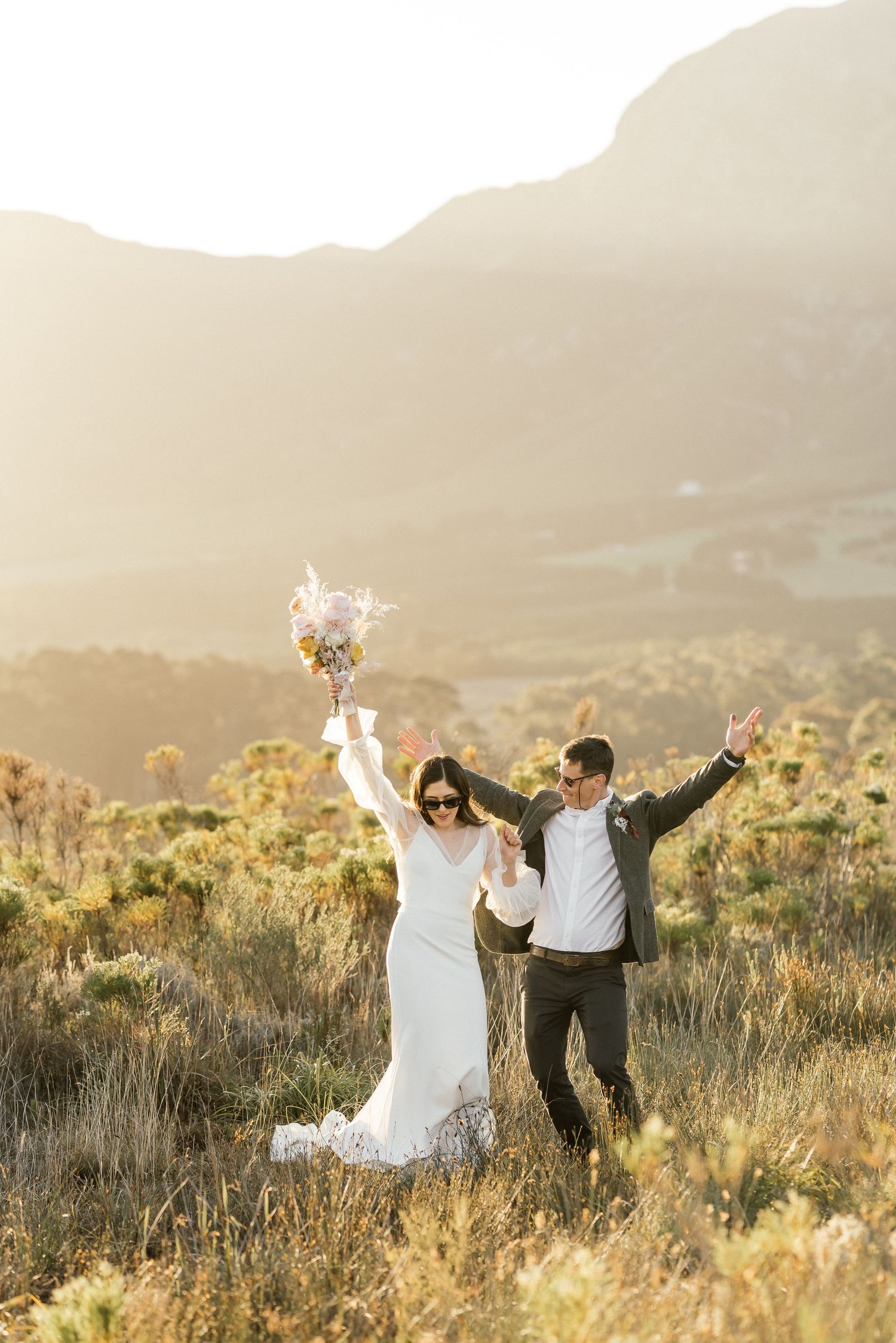 Bride and groom playfully dancing in sunglasses in a sunny field, with the bride raising her bouquet in celebration.