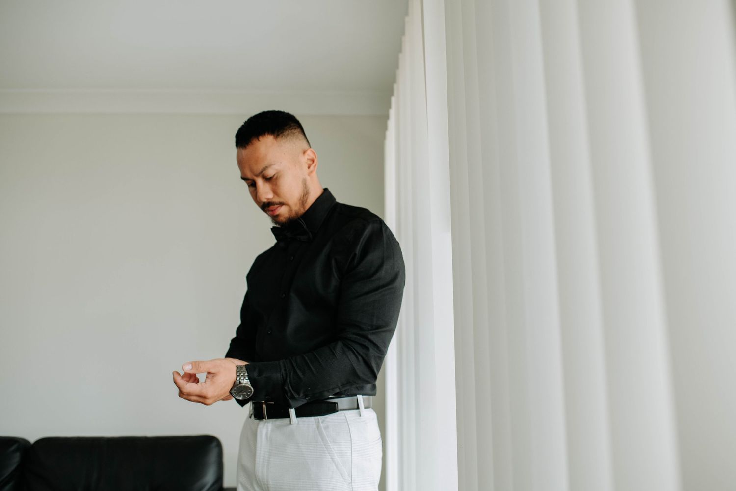 Person in black formal attire checking watch while standing near white curtains.