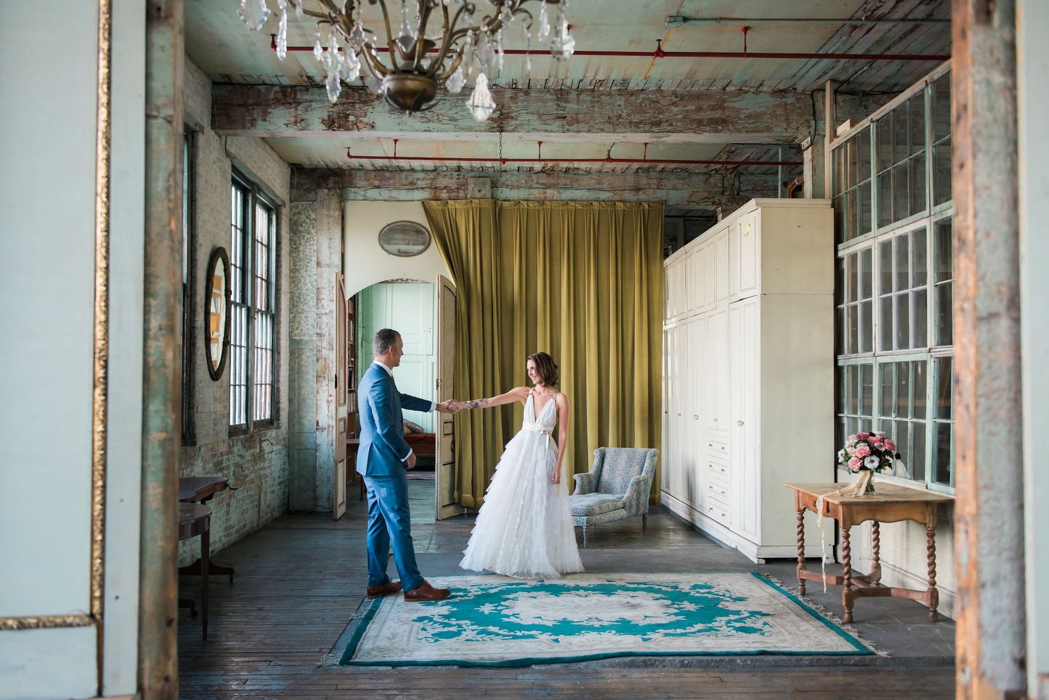 Two people stand together in their formal attire in an industrial loft space with rustic exposed walls and yellow curtains.