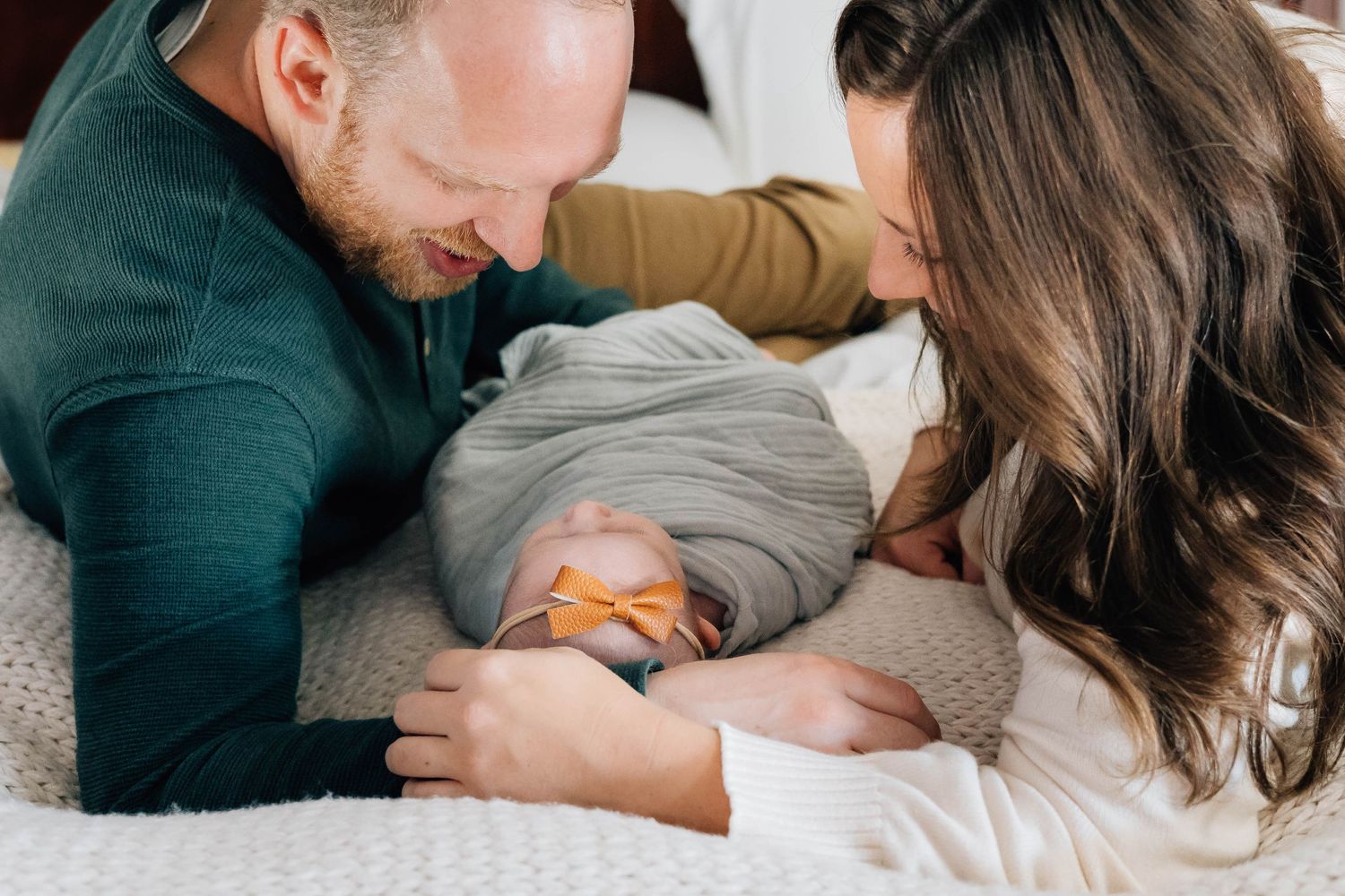 Two parents lying on a bed, smiling and looking down at their newborn baby together.