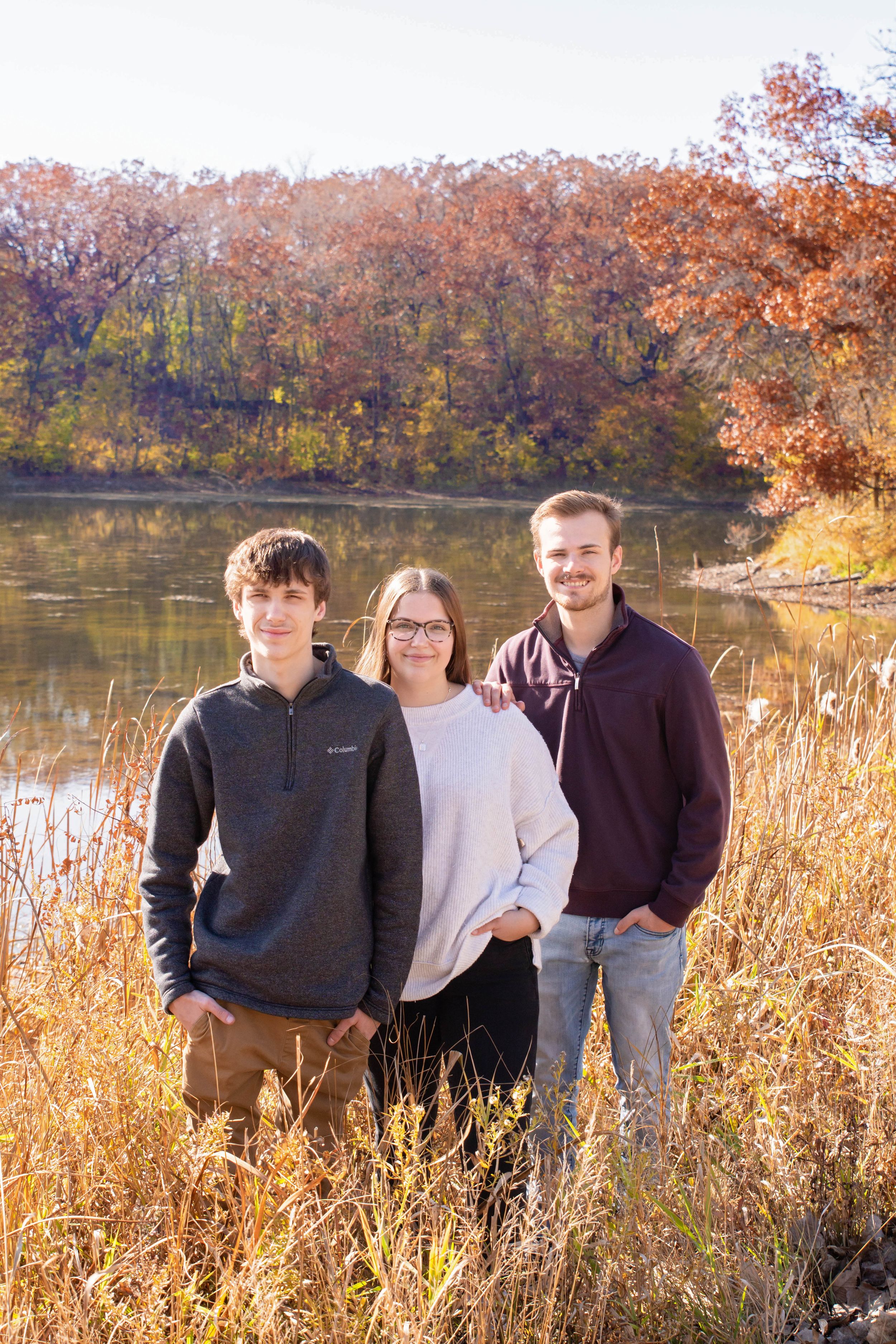 Three people standing together in tall autumn grass with fall foliage and a lake in the background.