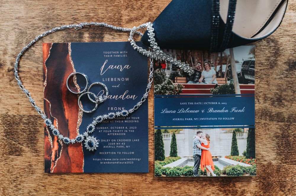 Navy blue wedding invitations with rings and jewelry on wooden surface.