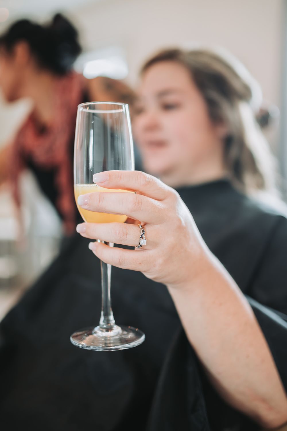 A hand holding a champagne flute with a diamond ring visible during wedding preparations.