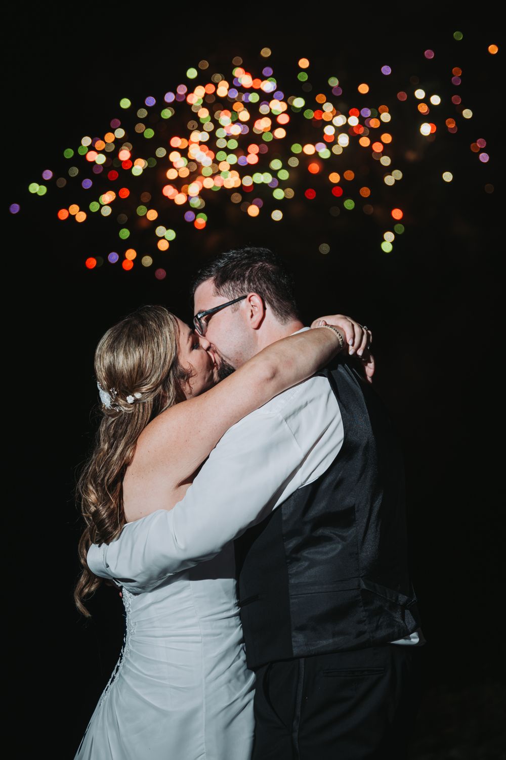 A couple shares their first dance as newlyweds beneath colorful bokeh lights in the night sky.