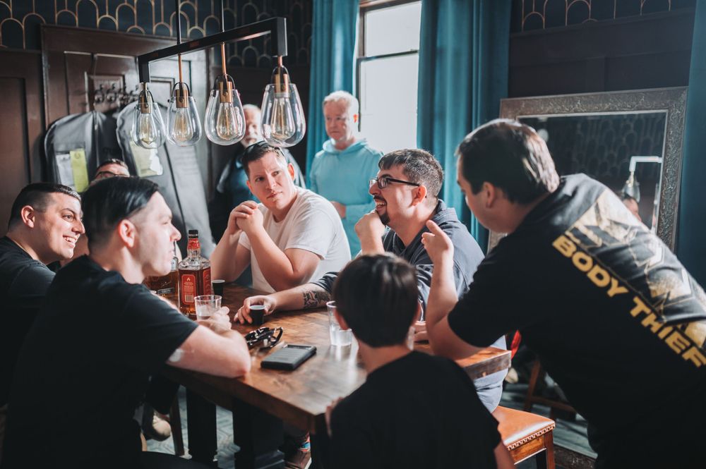 A group relaxes with drinks at a casual gathering around a table in a dimly lit setting.