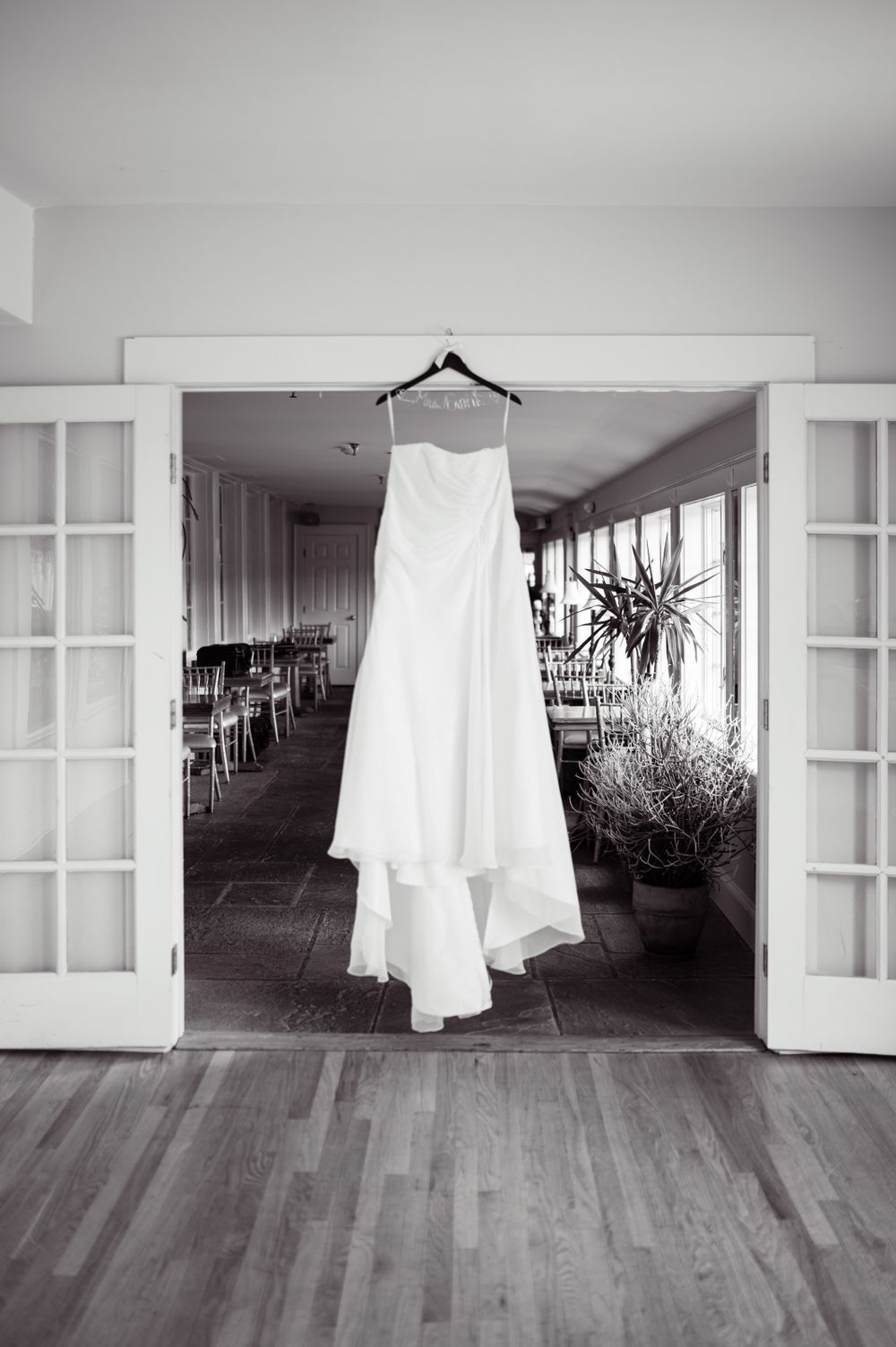 An elegant white wedding gown hangs in a doorway of a bright indoor venue with French doors.