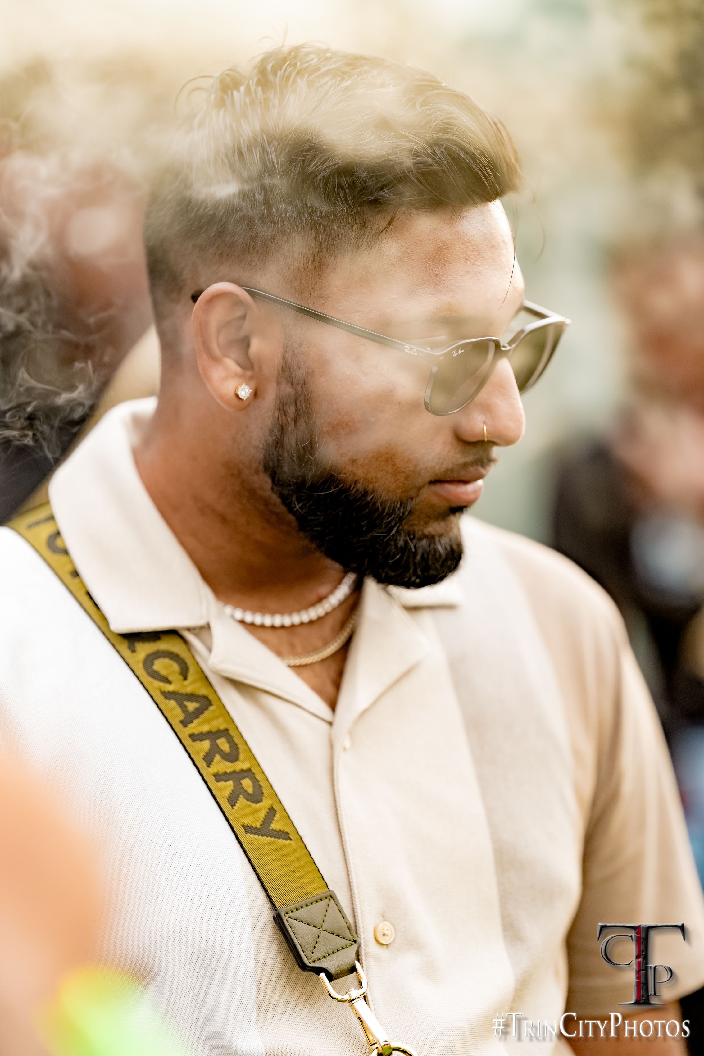 Man with beard, sunglasses, pearl necklace, and yellow GARRY strap poses outdoors at a city event.