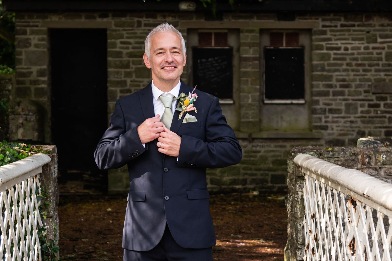 Happy couple poses on white wooden bridge in brick garden setting during wedding celebration.