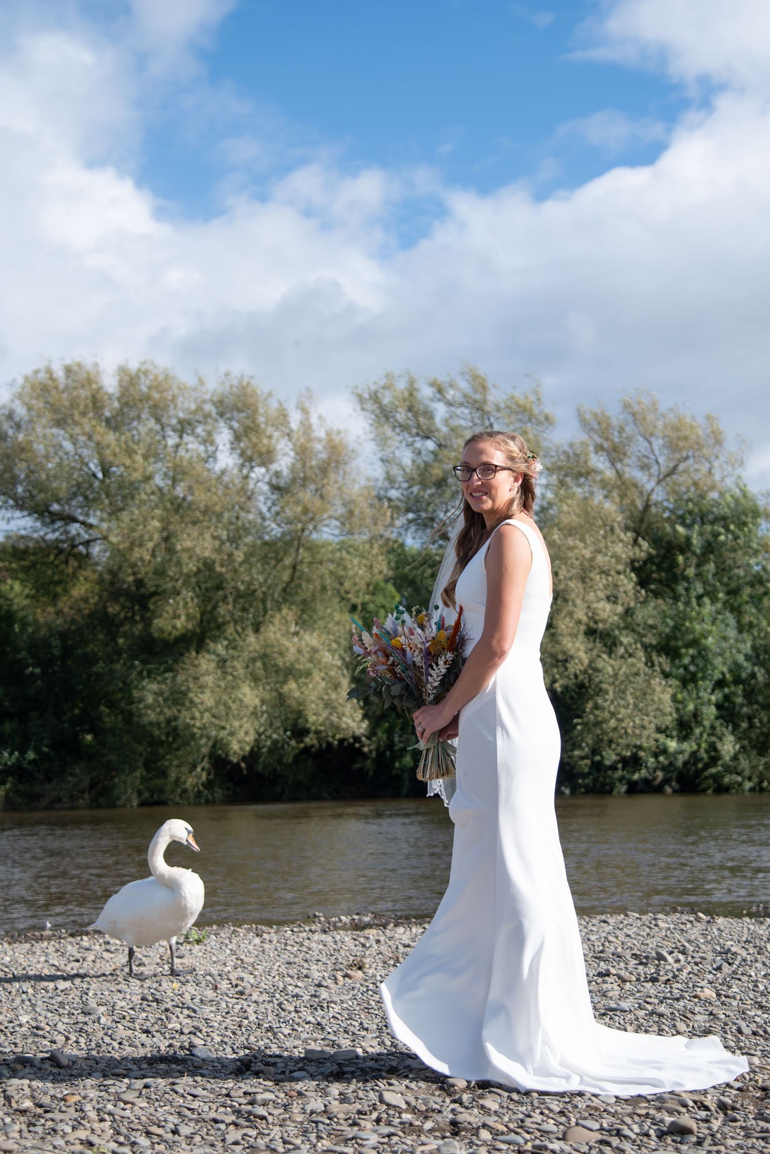 Elegant bride in white dress walks along rocky riverbank with swan in background.