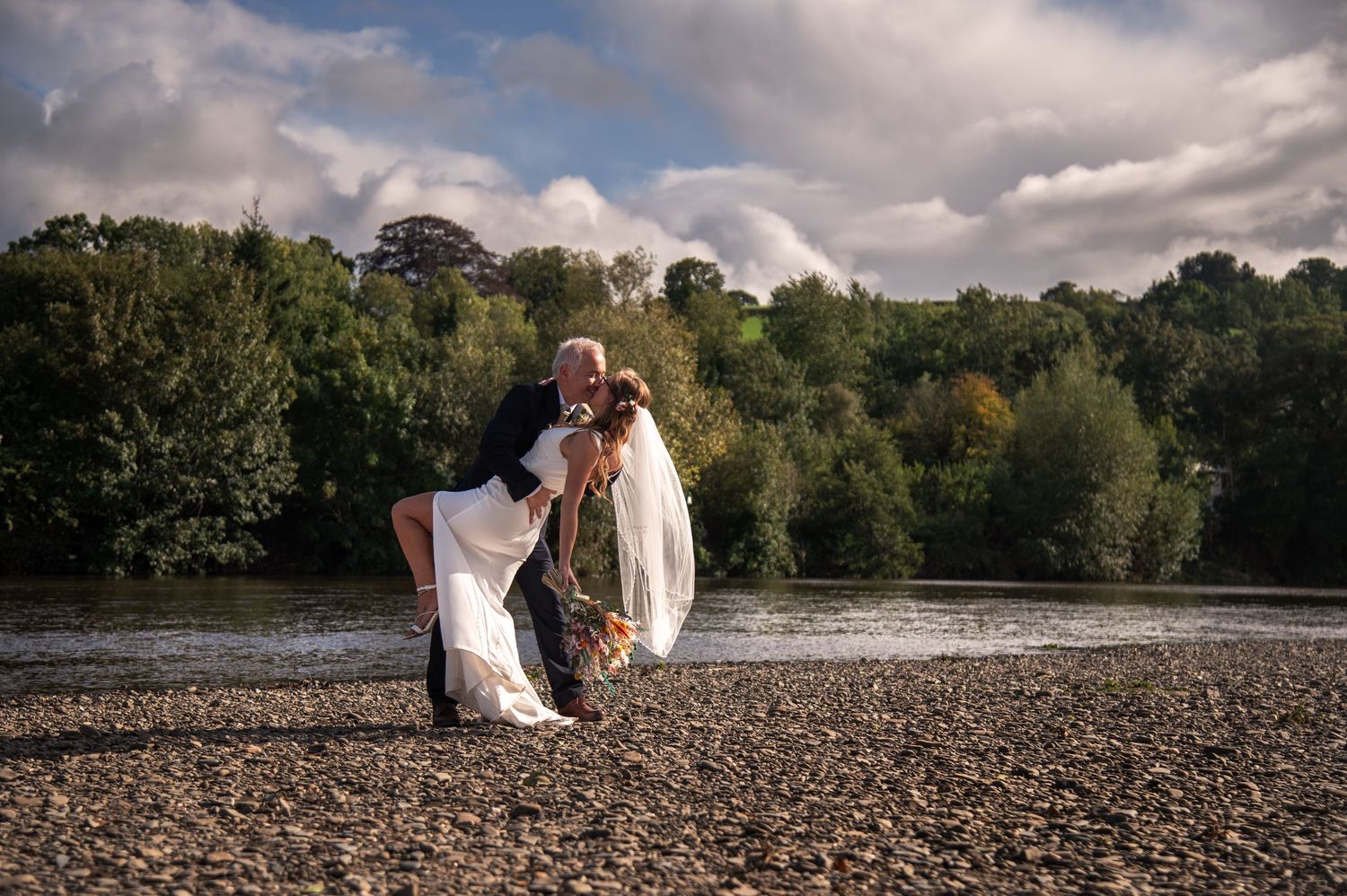 Couple poses for romantic photos near a scenic riverbank on a cloudy day.