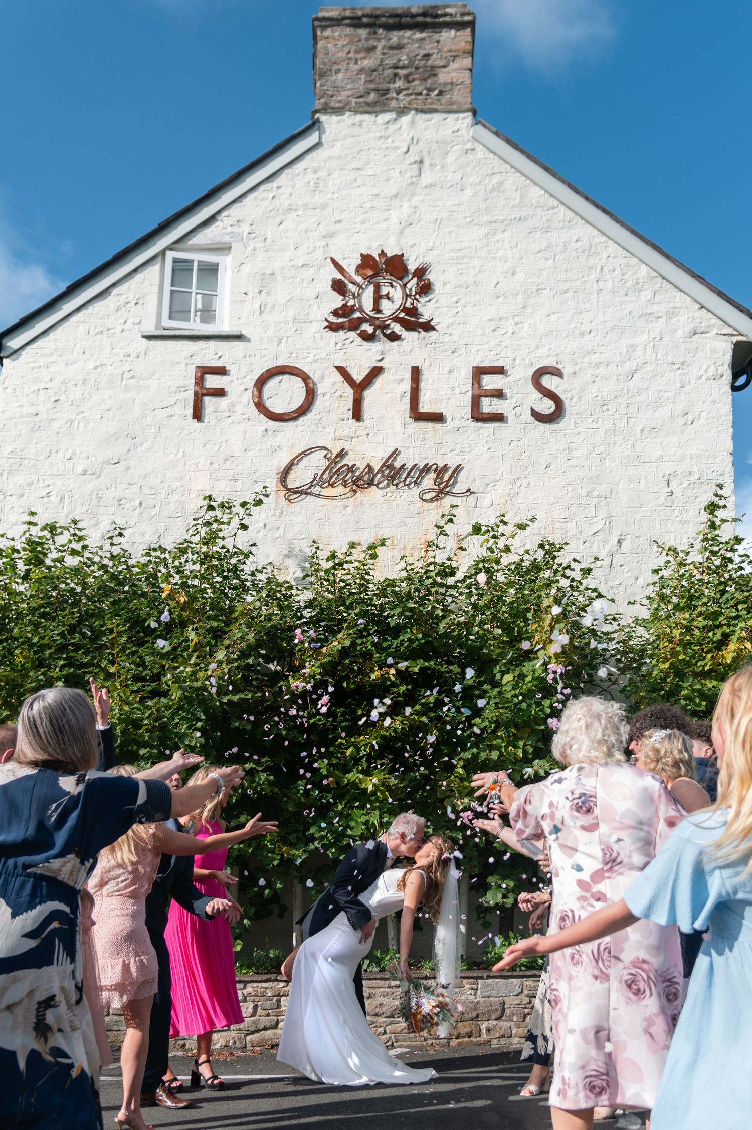 Wedding celebration outside white building with Foyles signage and flowering bushes.
