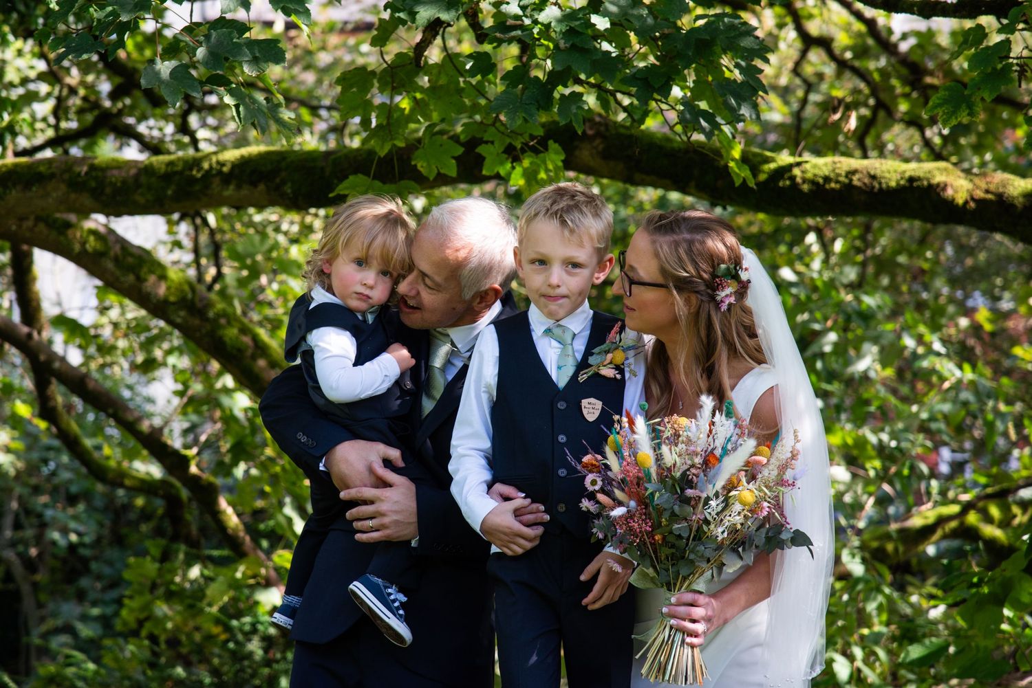 Wedding party poses outdoors under lush green trees on a sunny day.