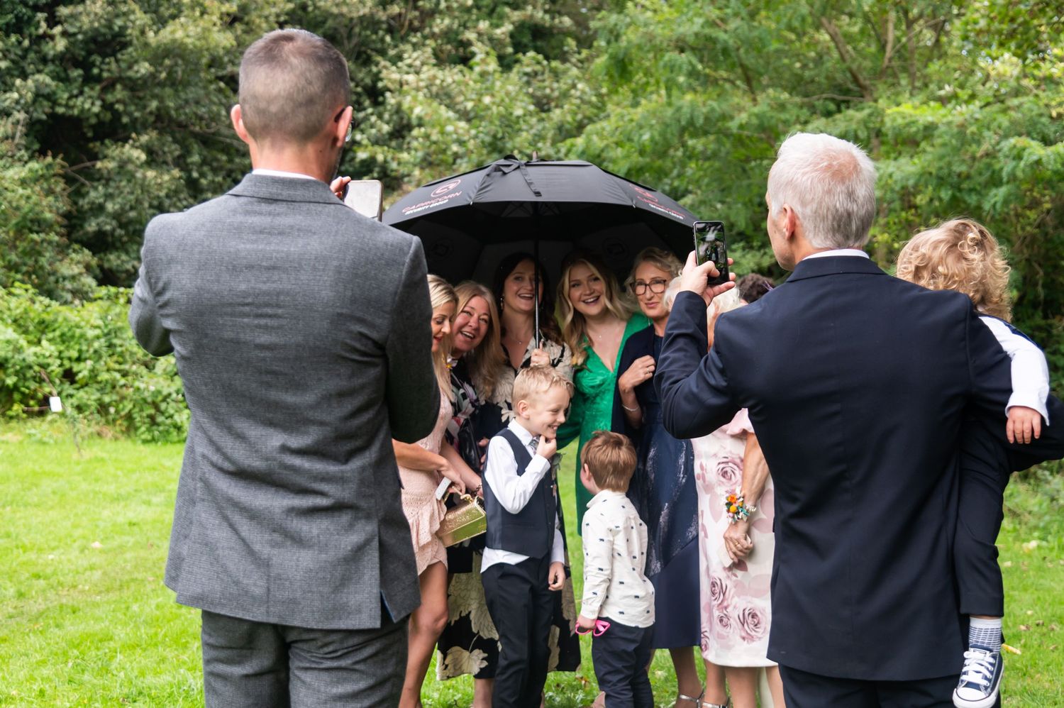 Wedding guests gather under umbrellas on a rainy day outdoors.