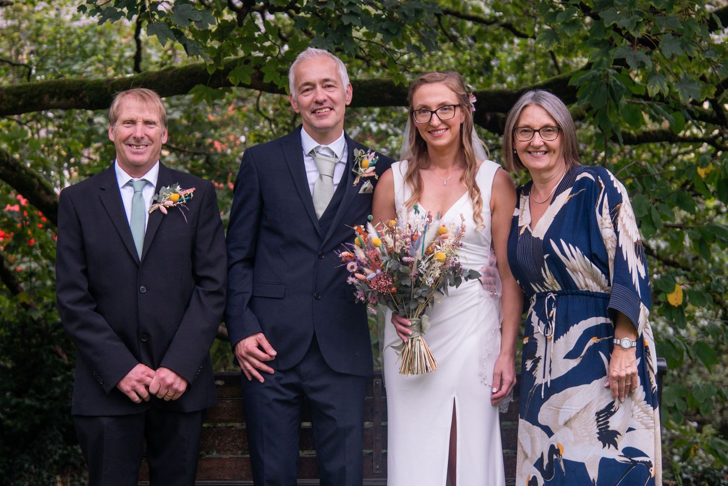 A wedding party poses together outdoors with greenery in the background.