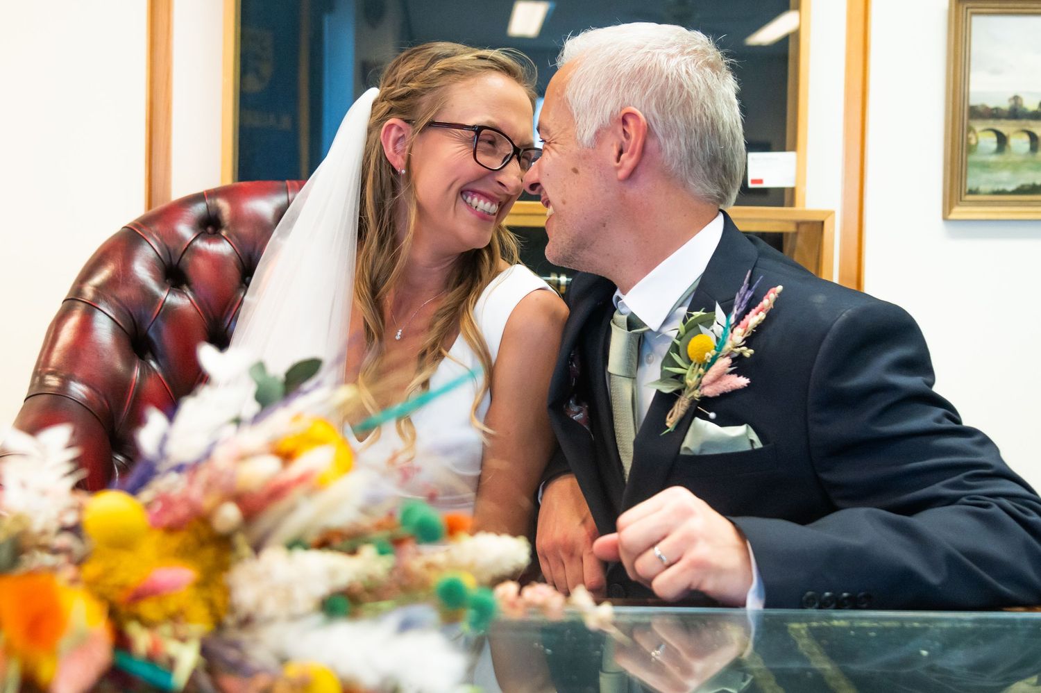 Wedding couple leaning in close to each other while sitting at a table with floral decorations.