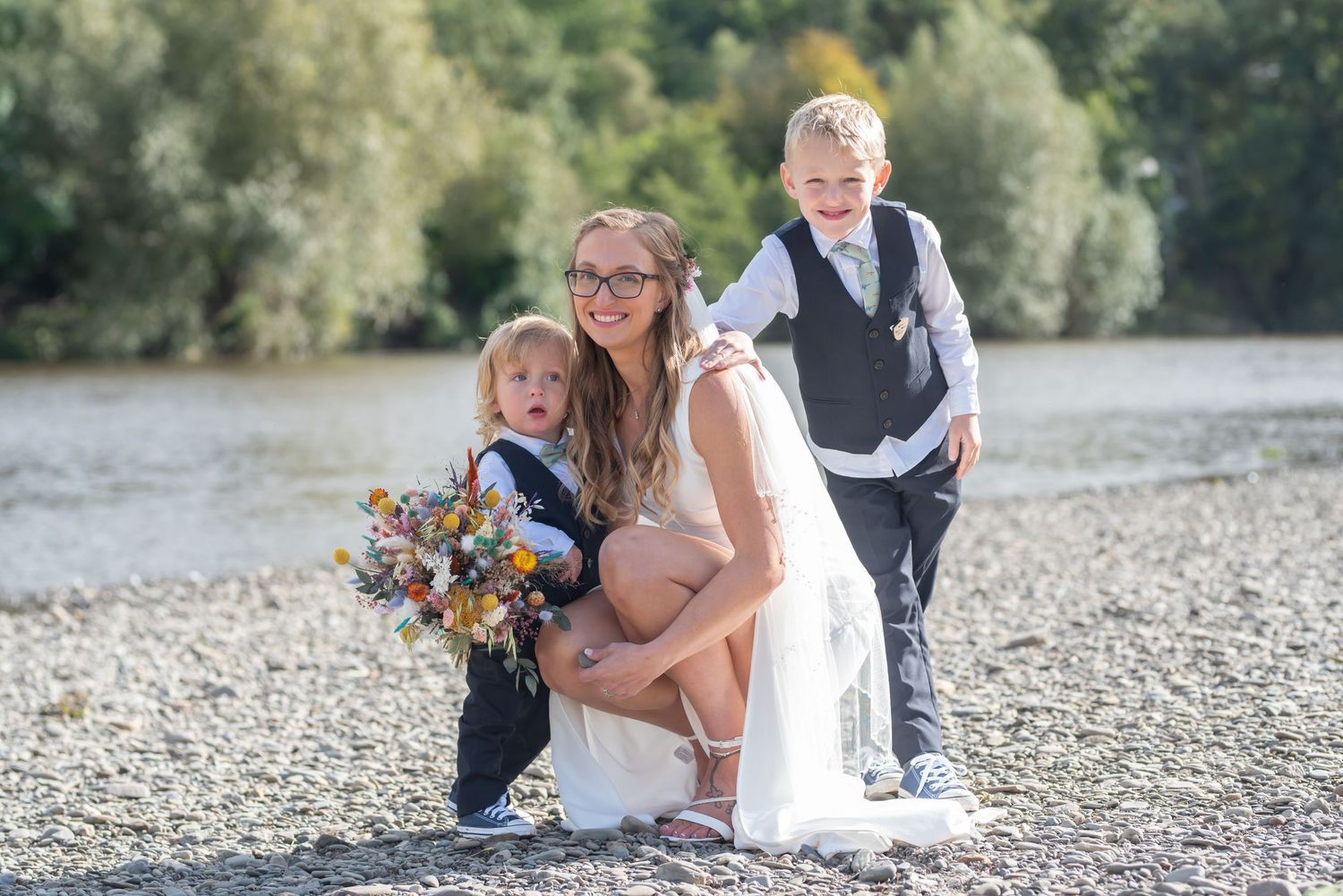 Wedding group relaxes on a rocky riverbank surrounded by trees.