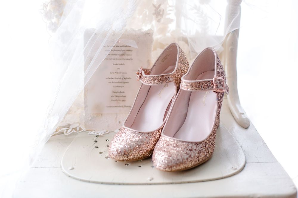 Rose gold glitter bridal heels displayed with wedding invitation and lace veil on white surface.