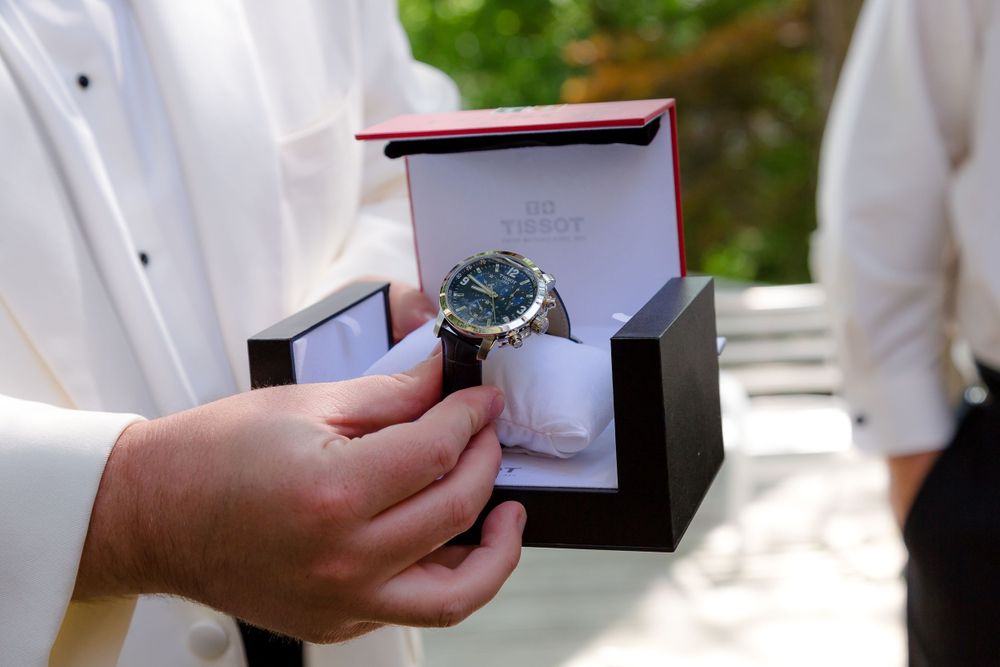Groom holding open red Tissot watch box as wedding gift during outdoor ceremony.