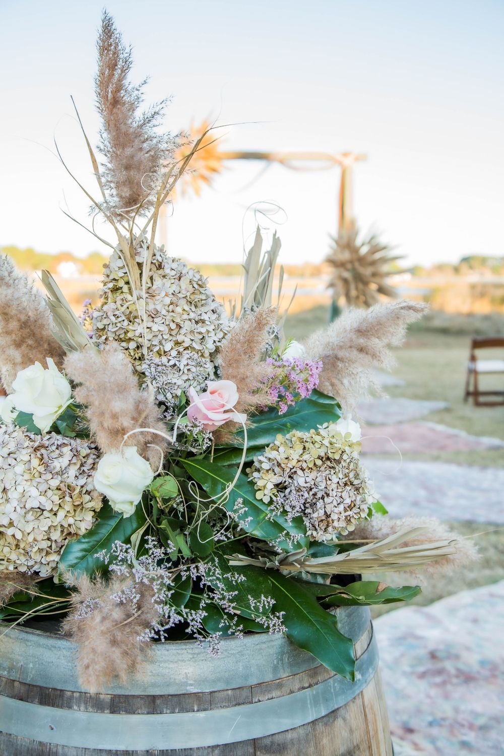 Rustic boho wedding ceremony decor with pampas grass, hydrangeas, and pink roses on a wine barrel.