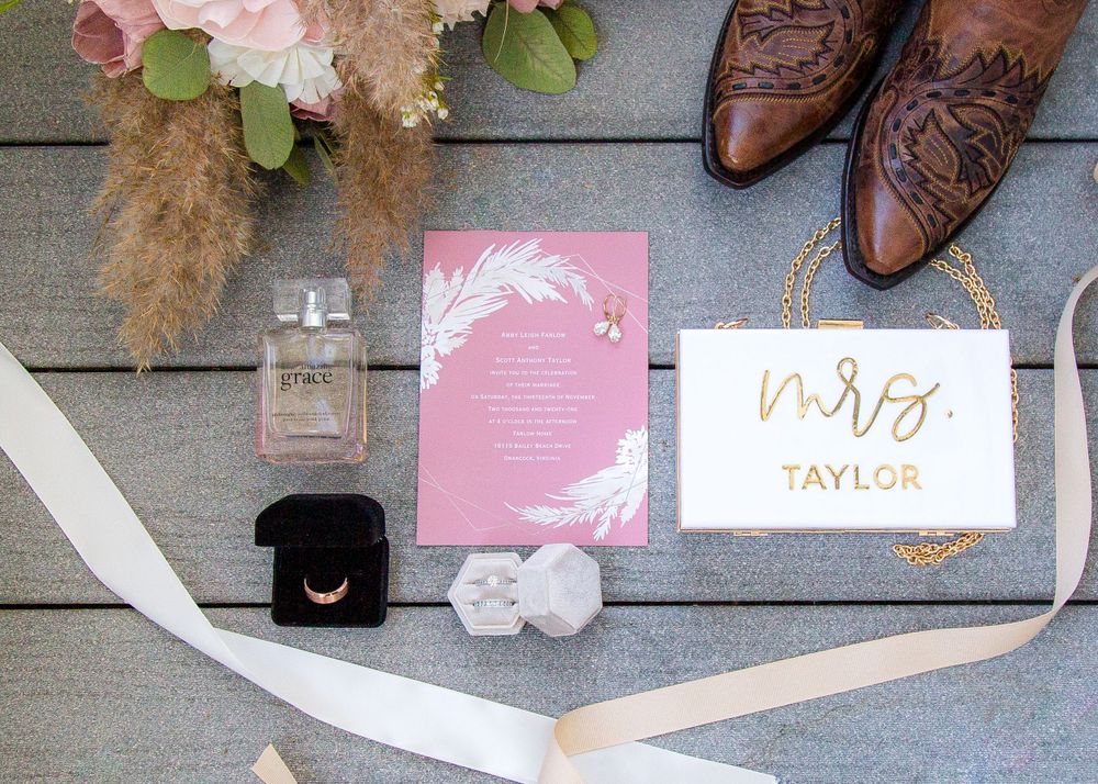 Pink and gold wedding invitation flat lay with perfume, ring box, boots, and bridal bouquet.