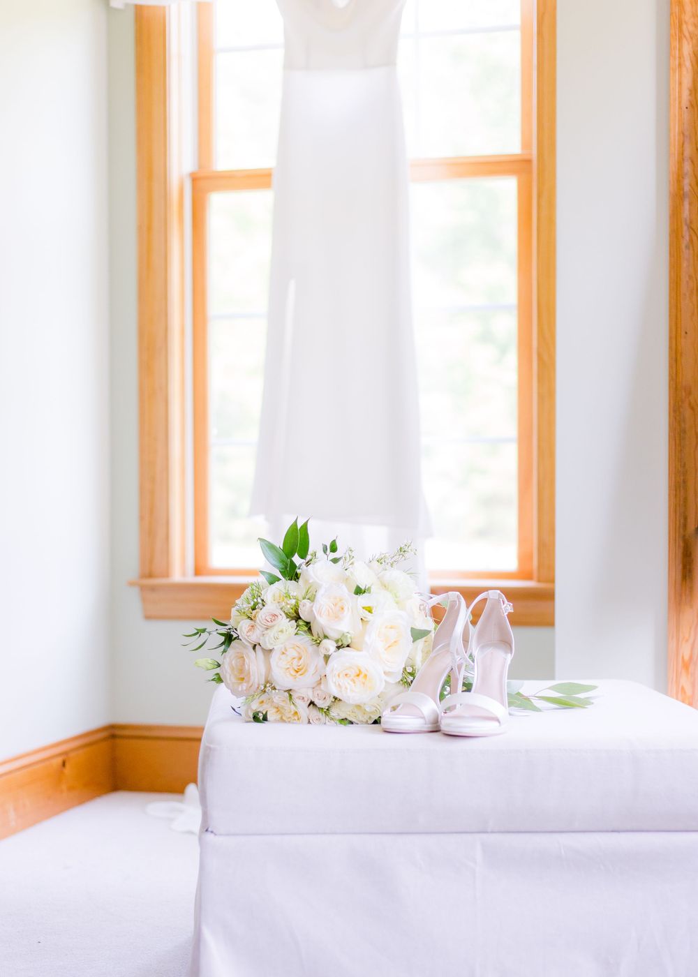 White wedding dress hanging by window with white rose bouquet and bridal heels on bench.