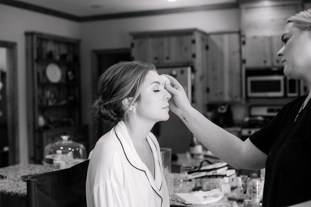 Black and white photo of bride getting makeup applied by artist in kitchen before wedding.