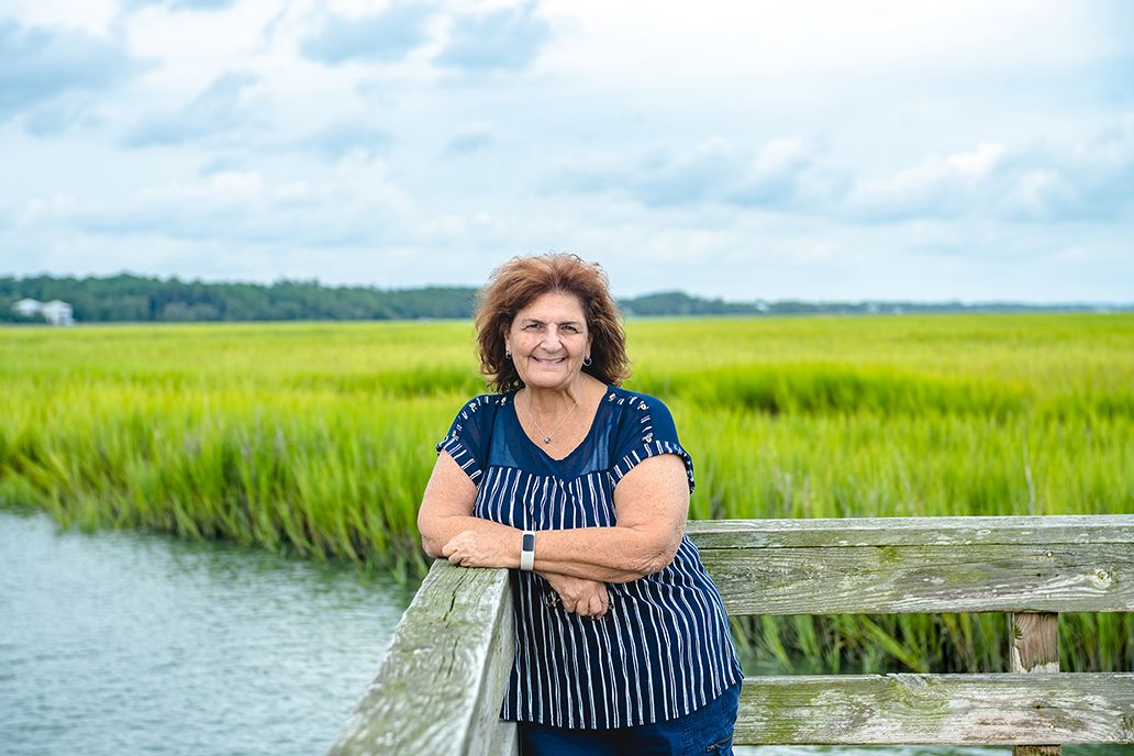A woman posing on a wooden dock overlooking a green salt marsh landscape.