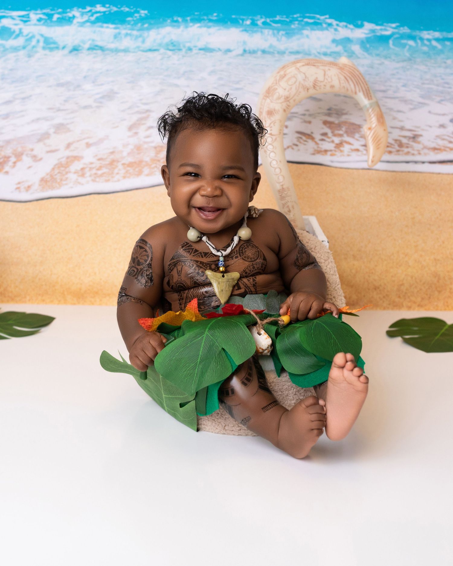 A happy baby sits on a beach backdrop with tropical leaves and ocean waves.