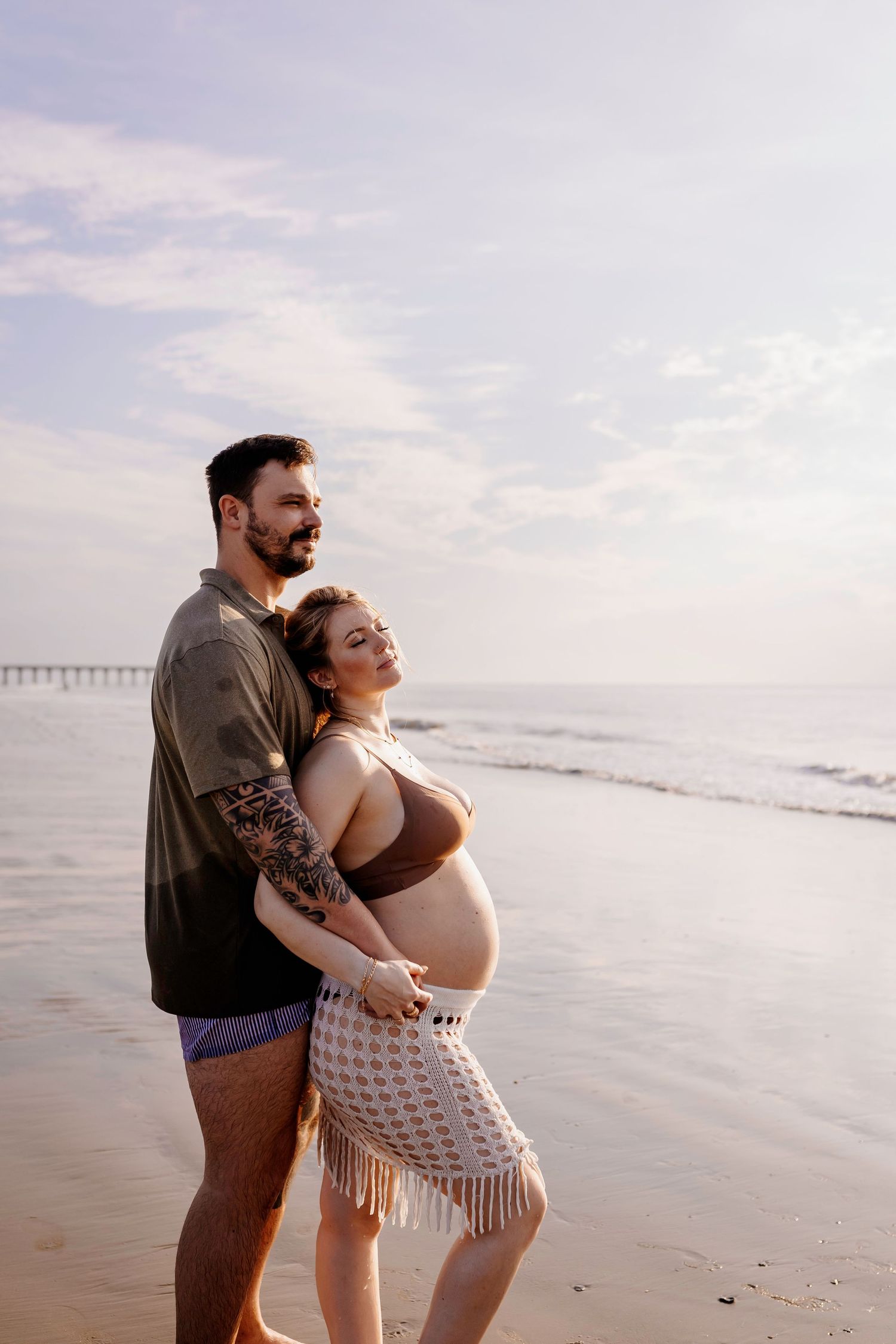 An expecting couple embraces on the beach with a pier visible in the background.