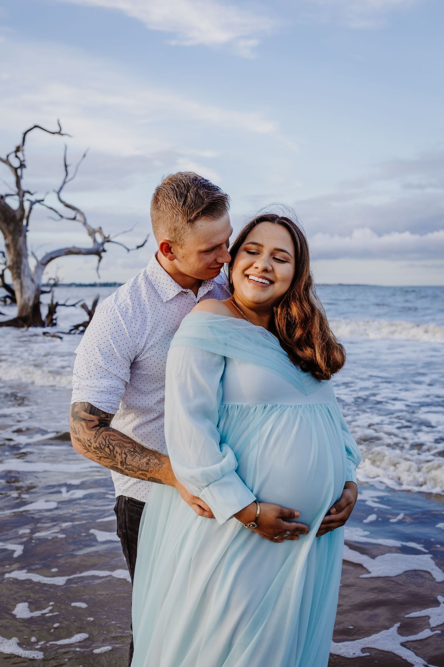A couple shares an intimate moment during their maternity photoshoot on a beach at sunset.