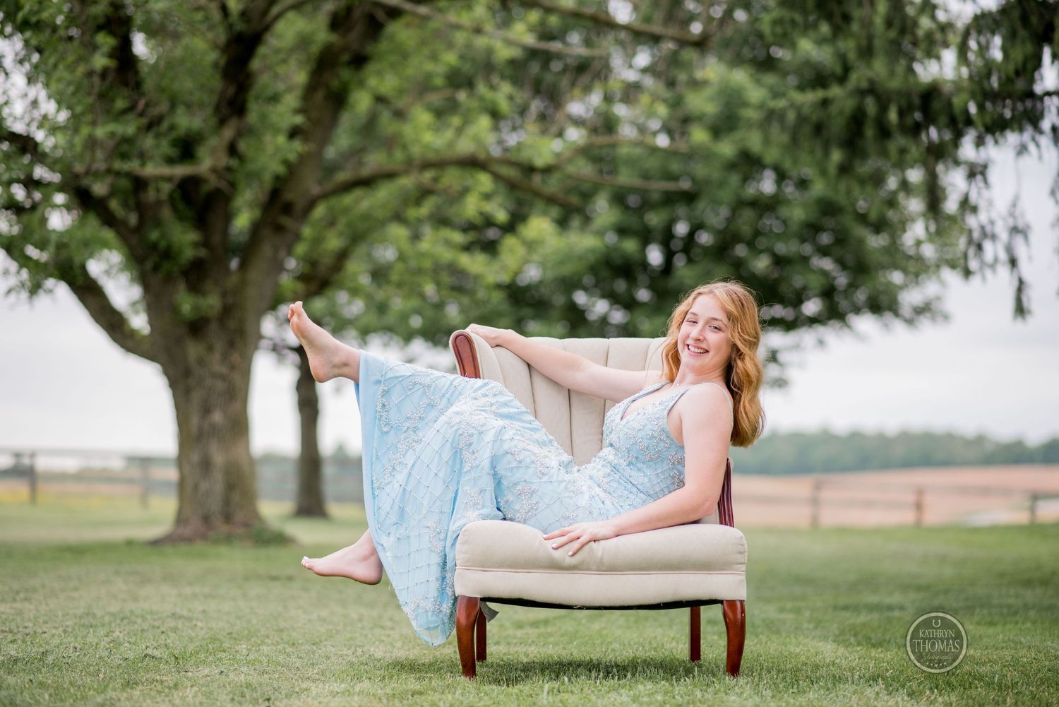Woman in a blue gown reclines playfully across a beige armchair on a grassy lawn.